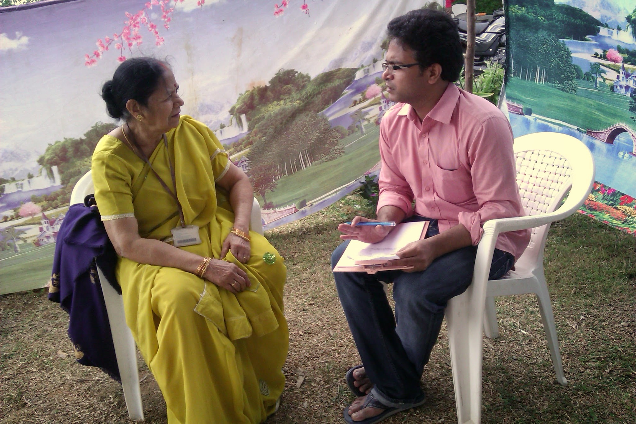 Dr. Suresh Kumar sitting on a plastic chair on the grass with a clipboard, listening one-on-one with humility to an elderly mother